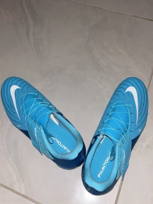 Nike Phantom GX blå fotbollsskor - Säljer ett par Nike Phantom GX fotbollsskor i en snygg blå färg med vita detaljer och klassisk Nike-logga. Skorna har snörning och kardborreband för extra bra passform. Tillverkade i syntetmaterial som ger bra bollkontroll och komfort på planen. Säljer pågrund av de är för stora