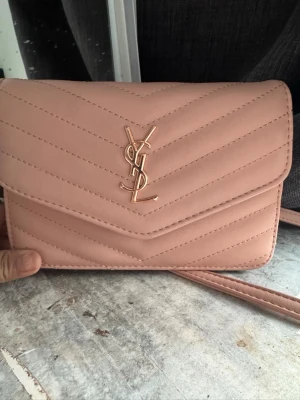 Ljusrosa YSL quiltad handväska - Supersnygg ljusrosa handväska från YSL med quiltat mönster och guldfärgad logga framtill. Väskan har en axelrem i matchande färg och en stilren, rektangulär form. Perfekt för dig som vill ha en trendig accessoar med lyxig känsla.