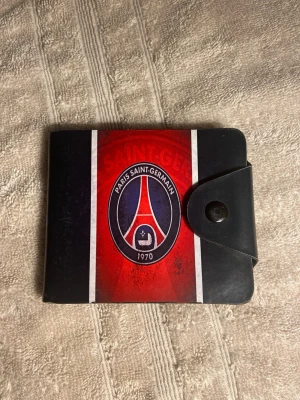 PSG plånbok i svart och rött - Snygg plånbok med Paris Saint-Germain-logga i rött, svart och vitt. Plånboken har tryckknapp, flera kortfack och är gjord i konstläder. Perfekt för dig som älskar fotboll och vill visa vilket lag du hejar på.