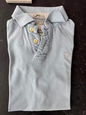 Ljusblå piké från Abercrombie & Fitch - Säljer en ljusblå pikétröja från Abercrombie & Fitch med korta ärmar och klassisk krage. Tröjan har två gula knappar och en broderad älglogga på bröstet. Tillverkad i mjuk bomull för en bekväm känsla.