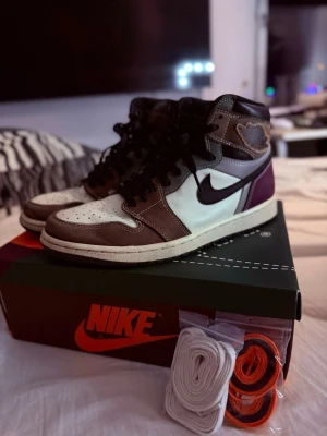 Nike Air Jordan 1 High Mocha - Säljer ett par Nike Air Jordan 1 High Mocha sneakers med svart swoosh, bruna och vita paneler samt lila detaljer vid hälen. Skorna har högt skaft och kommer med extra skosnören i vitt och orange. Klassisk AJ1-design i läder och mocka.