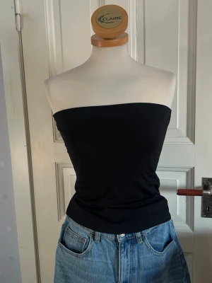 Svart bandeau tubtopp - En enkel och stilren svart bandeau tubtopp som sitter tight runt överkroppen. Toppen är axelbandslös och passar perfekt till jeans eller kjol för en clean look. Materialet känns mjukt och stretchigt, vilket gör den bekväm att bära hela dagen.