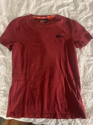 Röd Superdry t-shirt i bomull - Snygg röd t-shirt från Superdry med broderad logga på bröstet. Klassisk passform och rund halsringning. Tillverkad i mjuk bomull som känns skön mot huden. Perfekt för en avslappnad och stilren look.