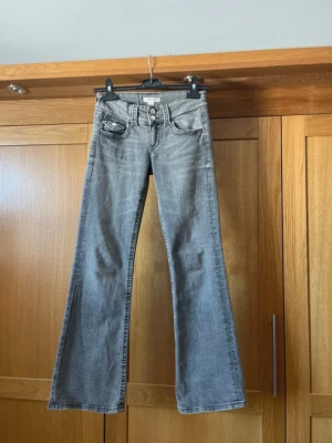 Grå bootcut jeans från Gina tricot - Säljer ett par grå bootcut jeans från gina tricot med dubbla knappar i midjan och en unik fickdetalj med lock och knapp. Jeansen har en tvättad look och klassisk femficksdesign. Perfekta för dig som gillar retrostil och vill ha något som sticker ut.