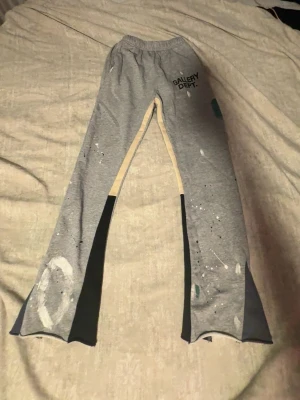 Grå mjukisbyxor Gallery Dept. - Unika grå mjukisbyxor från Gallery Dept. med svart och beige kontrastpaneler längs benen och färgstänk som ger en kreativ vibe. Byxorna har resår i midjan och utsvängda ben. Perfekta för dig som gillar streetwear och vill sticka ut.