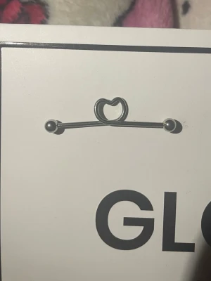 Industriell stav med hjärtdetalj - Säljer en silvrig industriell stav i metall med en söt hjärtformad detalj i mitten. Båda ändarna har kulor som skruvas av för enkel användning. Perfekt för dig som vill ha en unik och stilren piercingaccessoar.