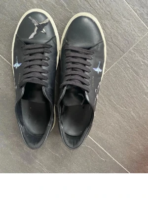 Svarta Axel Arigato sneakers med fågelmotiv - Snygga svarta sneakers från Axel Arigato med färgglada fågelmotiv på sidorna och ovandelen. Skorna har svarta skosnören, vit sula och är tillverkade i läder. Perfekta för dig som vill ha en unik och trendig look.