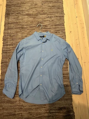 Blå skjorta från Polo Ralph Lauren - Klassisk ljusblå skjorta från Polo Ralph Lauren med button-down krage och gul broderad logga på bröstet. Skjortan har lång ärm, knappar framtill och är tillverkad i mjuk bomull. Perfekt för en clean och stilren look. Knappt använd. L men funkar på M
