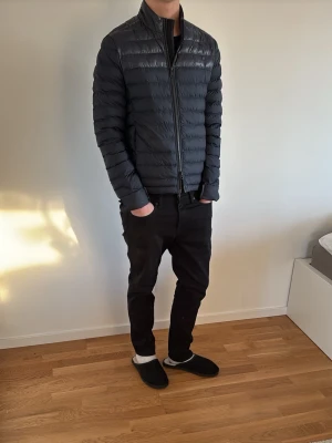 Svart dunjacka från J.Lindeberg - Snygg svart dunjacka från J.Lindeberg med quiltad design och hög krage. Jackan har dragkedja framtill och två sidofickor. Perfekt för kalla vinterdagar och har en stilren, smal passform. Materialet är lätt och värmande dun.