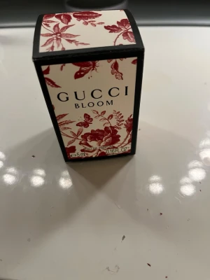 Gucci Bloom Eau de Parfum 5ml - Gucci Bloom Eau de Parfum i miniflaska på 5 ml. Flaskan är beige med svart-vit etikett och har en stilren, fyrkantig form. Ytterkartongen är dekorerad med röda blommor och blad mot vit bakgrund, vilket ger en lyxig och elegant känsla.