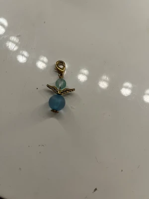Blå ängelberlock med guldfäste - Söt berlock formad som en ängel med två blå glaspärlor och guldfärgade detaljer. Har en guldfärgad karbinhake som gör det enkelt att fästa på armband, halsband eller väska. Perfekt accessoar för att ge din stil en personlig touch.
