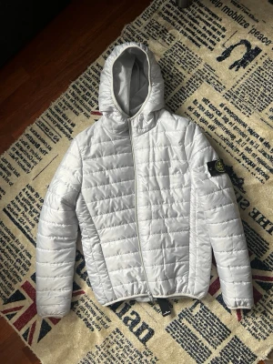 Vit pufferjacka från Stone Island - Säljer en stilren vit pufferjacka från Stone Island med ikonisk patch på vänster ärm. Jackan har huva, dragkedja framtill och quiltad design. Perfekt för kalla dagar och ger en clean look. Materialet är lätt och isolerande för extra värme.