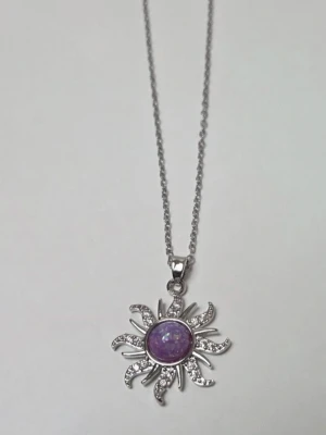 Silverhalsband med lila solhänge Rostfritt Stål  - Snyggt halsband i silverfärg med ett solformat hänge. Hänget har en lila, skimrande sten i mitten och är dekorerat med små gnistrande stenar på solstrålarna. Kedjan är tunn och justerbar med karbinlås. Perfekt för dig som gillar unika detaljer. Detaljer: Design: Celestial / Sol / Stjärna Material: Rostfritt stål (Stainless Steel) – tål vatten & missfärgas inte Stenar: Lila opal-liknande sten & gnistrande syntetisk zirkon Hänge: 2,2 cm Kedjelängd: 45 cm Skick: Nytt 