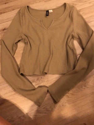 Beige ribbad croppad topp divided - Trendig beige croppad topp från divided med lång ärm och ribbad struktur. Toppen har en snygg v-ringad detalj vid halsen och är perfekt för att styla med höga jeans eller kjol. Enkel men ändå stylish design som passar till många looks.