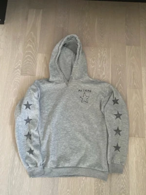 Grå hoodie med stjärnor och text - Snygg grå hoodie med huva och dragsko. Svarta stjärnor längs båda ärmarna och texten 'MA CHERIE' med en stjärna på bröstet. Mjuk och bekväm, perfekt för chill dagar. Tillverkad i bomullsblandning.