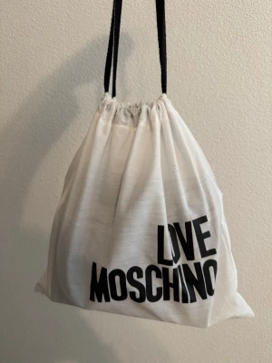 Love Moschino svart axelväska - Snygg svart axelväska från Love Moschino med justerbar axelrem och guldfärgad kedjedetalj framtill såg går att ta av ifall man vill. Dustbagen tillkommer 💕