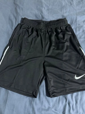 Svarta träningsshorts från Nike - Säljer ett par svarta träningsshorts från Nike med elastisk midja och vit Nike-logga på benet. Shortsen har en sportig look med vita detaljer på sidorna och är tillverkade i ett lätt och snabbtorkande material.