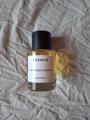 ESSNCE Chocolat Chaud Eau de Parfum - ESSNCE Chocolat Chaud Eau de Parfum kommer i en rund glasflaska med svart lock och enkel vit etikett. Doften har en varm, chokladig ton och flaskan har en stilren, minimalistisk design. Perfekt för dig som gillar söta och mysiga dofter.