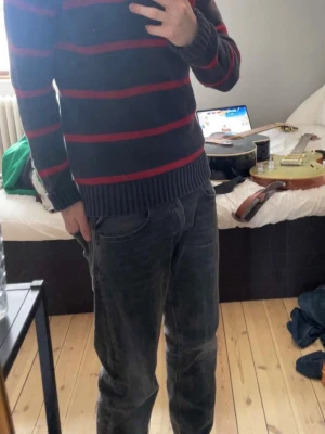 Randig stickad tröja från Polo Ralph Lauren -  Mörkblå stickad tröja med röda ränder från Polo Ralph Lauren. Tröjan har halv dragkedja vid halsen, ribbade muddar och nederkant samt den klassiska broderade loggan på bröstet. Perfekt för en avslappnad men snygg stil. Storlek L (14-16) passar xs-s.