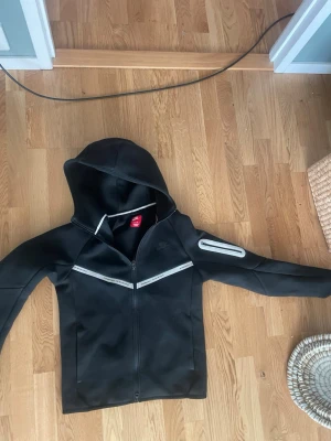 Svart hoodie från Nike med dragkedja - Snygg svart hoodie från Nike med dragkedja framtill och reflekterande detaljer på bröstet och ena ärmen. Hoodien har en klassisk huva och sportig look, perfekt för dig som gillar streetwear eller träningsstil.
