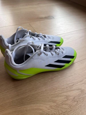 Adidas vita och neongröna inomhusskor - Säljer ett par snygga inomhusskor från Adidas i vitt med neongröna detaljer och svarta ränder på sidan. Skorna har låg profil, snörning och greppvänlig sula i gummi, perfekt för futsal eller inomhusträning.