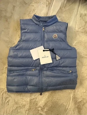 Ljusblå dunväst från Moncler - Snygg ljusblå dunväst från Moncler med hög krage och dragkedja framtill. Västen har två dragkedjeförsedda fickor och ikonisk Moncler-logga på bröstet. Tillverkad i glansigt material och fylld med dun för extra värme. Perfekt för lager-på-lager-stil.