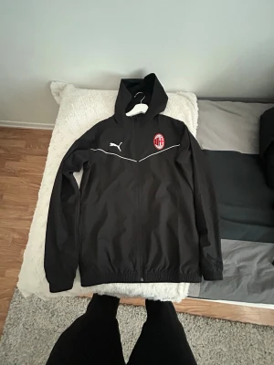 Svart AC Milan hoodie från Puma - Säljer en svart hoodie från Puma med AC Milan-logga på bröstet. Jackan har dragkedja, huva och vita detaljer längs bröstet samt Puma-logga. Perfekt för dig som gillar fotboll och vill ha en snygg och sportig look. Materialet är lätt och andas.