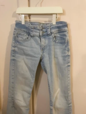 Ljusblå bootcut jeans med dubbla knappar - Säljer ett par ljusblå bootcut jeans med dubbla knappar i midjan och klassiska fem fickor. Jeansen har en snygg tvättad look. Perfekta för dig som gillar retrostil och vill ha ett par bekväma jeans i garderoben.