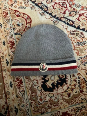 Grå Moncler mössa med ränder - Snygg grå stickad Moncler mössa med klassiska ränder i vitt, rött och marinblått vid kanten. Mössan har Moncler-logga framtill och är tillverkad i mjukt material som håller dig varm. Perfekt accessoar för kyliga dagar.