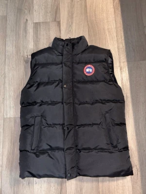 Svart dunväst från Canada Goose - Snygg svart dunväst från Canada Goose med hög krage och tryckknappar framtill. Västen har två fickor på sidorna och den klassiska Canada Goose-loggan på bröstet. Perfekt för lager-på-lager och håller dig varm under kyliga dagar.