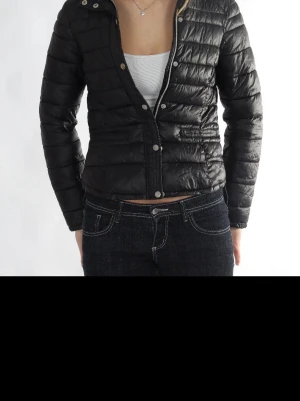 Svart kort pufferjacka - Snygg svart pufferjacka med kortare passform och quiltad design. Jackan har tryckknappar framtill och hög krage. Perfekt för kalla dagar och enkel att matcha med olika outfits. Materialet är glansigt och ger en modern look.
