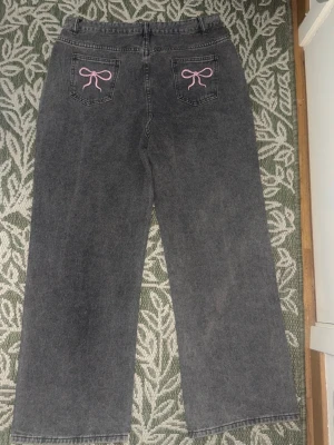 Gråa jeans med rosa rosetter - Säljer ett par gråa jeans med raka ben och broderade rosa rosetter på bakfickorna. Jeansen har hög midja, bälteshällor och klassisk femficksdesign. Perfekta för dig som vill ha en söt detalj på dina jeans!