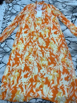 Orange blommig långklänning från Y.A.S - Snygg långklänning från Y.A.S i orange och vitt med blommigt mönster. Klänningen har lång ärm, v-ringning och är tillverkad i ett lätt, luftigt material som passar perfekt för varmare dagar.