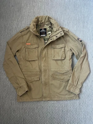 Beige vindjacka från Superdry - Stilren beige vindjacka från Superdry Rookie Edition med fyra framfickor, hög krage och camouflagemönstrat innerfoder. Jackan har detaljer som axelklaffar, tryckknappar och Superdry-logga på bröstet. Perfekt för en cool och avslappnad look.