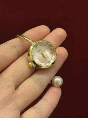 Guldig klocka med pärldetalj - Unik armbandsklocka med guldfärgat metallarmband och öppen design. Urtavlan är ljusrosa med små glittriga stenar som timmarkeringar. Avslutas med en stor pärldetalj på ena sidan för extra elegant touch.