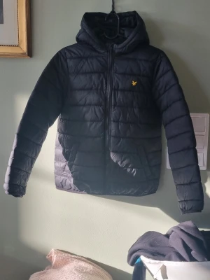 Svart pufferjacka från Lyle & Scott - Snygg svart pufferjacka från Lyle & Scott med gul broderad logga på bröstet. Jackan har huva, dragkedja framtill och två sidofickor. Tillverkad i ett glansigt, quiltat material som håller värmen bra under kalla dagar.