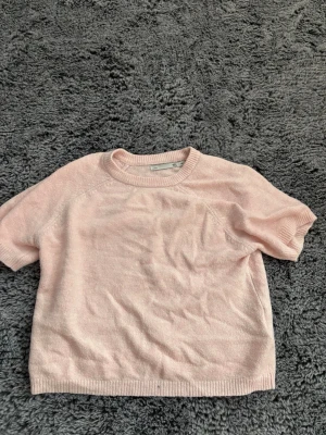 Ljusrosa stickad tröja  - Söt ljusrosa stickad tröja med korta ärmar och rund halsringning. Tröjan har ribbade muddar vid ärmslut och nederkant. Perfekt för lager på lager och en mjuk känsla. Materialet ser ut att vara bomull eller en bomullsblandning. Den är lite stor i storleken skulle jag säga.