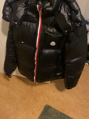 Svart dunjacka från Moncler - Snygg svart dunjacka från Moncler med glansig finish och klassisk Moncler-logga på bröstet. Jackan har en röd, vit och blå rand längs dragkedjan och stora vadderade partier som håller dig varm under vintern. Perfekt för dig som vill ha både stil och funktion. Äkta men har en lagning på bröstet därav priset