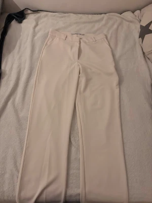 Vida beige kostymbyxor - Snygga beige kostymbyxor med låg midja och vida ben. Klassisk design med bälteshällor och pressveck framtill. Perfekta för en clean och stilren look. Materialet känns mjukt och har ett fint fall.