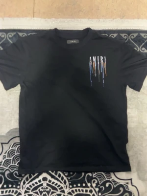 Svart AMIRI t-shirt med färgstänk - Snygg svart t-shirt från AMIRI med tryck på bröstet och ryggen. AMIRI-loggan är dekorerad med färgstänk i blått, vitt och orange som rinner ner för en cool streetwear-look. T-shirten har rund hals och korta ärmar, perfekt för dig som gillar trendiga detaljer.