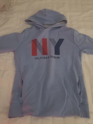 Ljusblå Hilfiger Denim hoodie - Snygg ljusblå hoodie från Hilfiger Denim med stor huva och tryck framtill med texten 'NY' i rött, vitt och blått. Mjuk bomullskänsla och breda muddar vid ärmslut och nederkant. Perfekt för en chill och avslappnad stil.