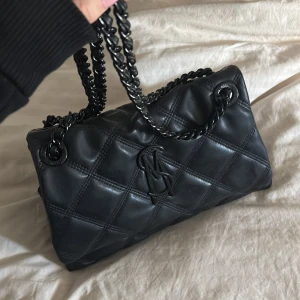 Svart quiltad handväska Steve Madden - Snygg svart quiltad handväska från Steve Madden med kedjehandtag i metall och diskret logga framtill. Väskan har en mjuk, puffig look och är tillverkad i konstläder. Perfekt för dig som gillar trendiga accessoarer med en lyxig känsla.