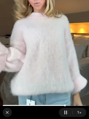 Ljusrosa fluffig stickad tröja - Supersöt ljusrosa stickad tröja med oversized passform och breda ärmar. Tröjan har en hög, ribbad halsringning och är gjord i ett mjukt, fluffigt material som ger en mysig känsla. Perfekt att styla med jeans för en trendig look.