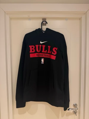 Bulls hoodie NBA Dri-FIT - Snygg svart hoodie från Nike med stor röd BULLS-text och NBA-logga på bröstet. Tillverkad i Dri-FIT-material som andas. Klassisk huva och känguruficka framtill. Perfekt för basketfans som vill ha en sportig och bekväm stil.                                                    Modellen är 185cm lång och väger 82kg.
