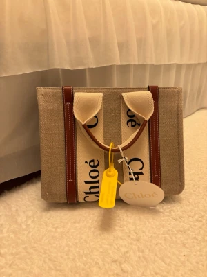 Chloé Woody beige handväska - Säljer en snygg beige handväska från Chloé i canvas med bruna läderdetaljer och vita handtag med Chloé-logga. Väskan är sprillans ny och helt oanvänd. 