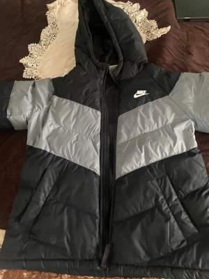 Nike svart och grå pufferjacka - Snygg svart och grå pufferjacka från Nike med huva och dragkedja framtill. Jackan har två sidofickor och ett stilrent mönster med diagonala färgblock. Perfekt för kalla dagar och har en sportig vibe med Nike-logga på bröstet.