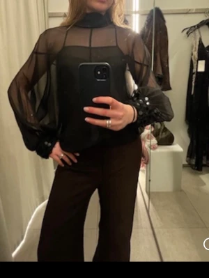 Svart blus  - Säljer en svart transparent blus med polokrage och långa, vida ärmar. Blusen har knappar vid ärmsluten och är riktigt flowy och snygg att styla lager på lager. Perfekt för dig som gillar statement-plagg och vill sticka ut lite extra. ( Helt ny, använd en gång ) 