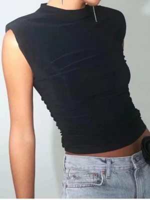 Croppad topp med rynkade sidor - Snygg svart croppad topp med höga axlar och rynkade sidor. Toppen är ärmlös och har en tight passform som framhäver figuren. Färgen är mörkgrå.
