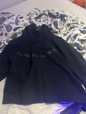 Svart Nike hoodie med dragkedja - Svart hoodie från Nike med hel dragkedja framtill och två fickor. Snygg detalj med en svart rand över bröstet. Klassisk huva och diskret Nike-logga på vänster sida. Perfekt för en sportig och avslappnad stil.