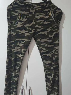 Camo leggings med dragkedjor - Snygga leggings med camouflage-mönster i grönt, svart och beige. De har två fejkade fickor framtill med guldfärgade dragkedjor och bred resår i midjan för skön passform. Perfekta för en avslappnad och trendig look.